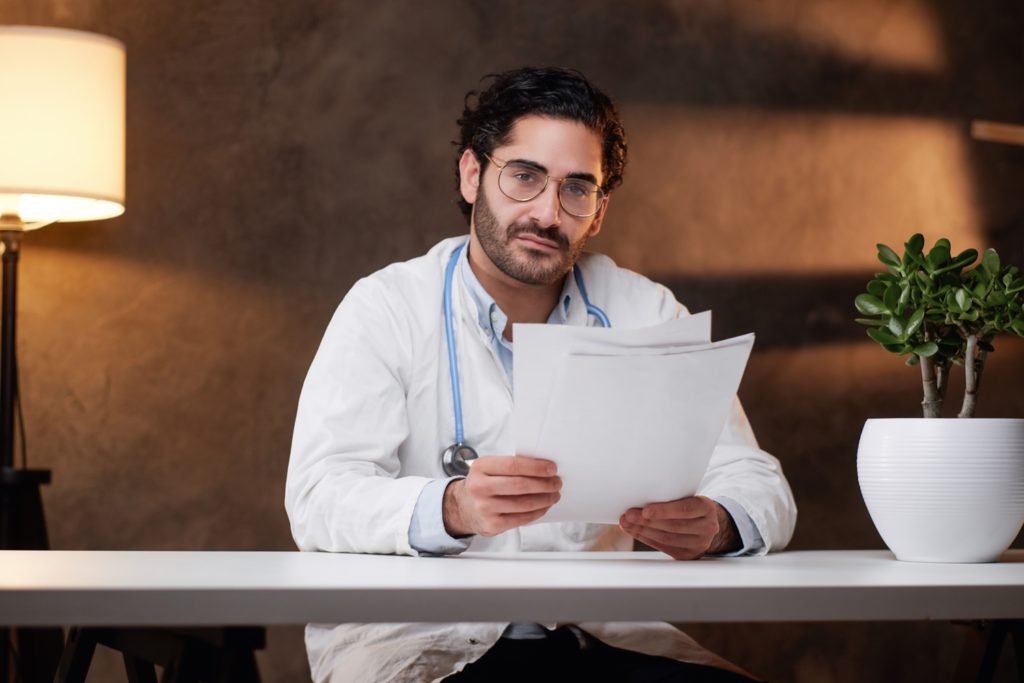 A Doctor Specialist Sitting At The Table Holding Papers In Room - SQUIPP CONSULTORIA E ASSESSORIA CONTÁBIL